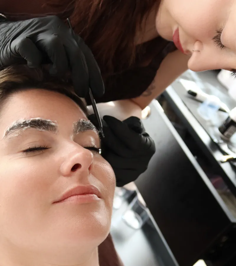 Gloved hands tweezing stray hairs to tidy a client's brows before treatment