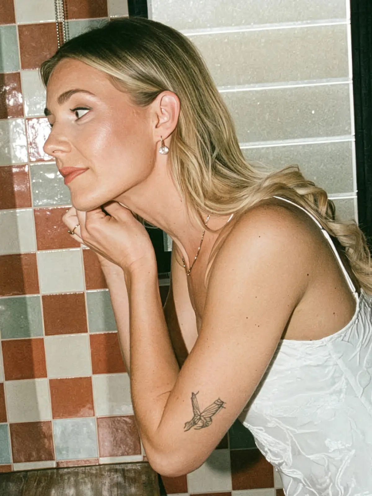 Bride with glowing, natural makeup and soft blonde waves, leaning against a tiled wall