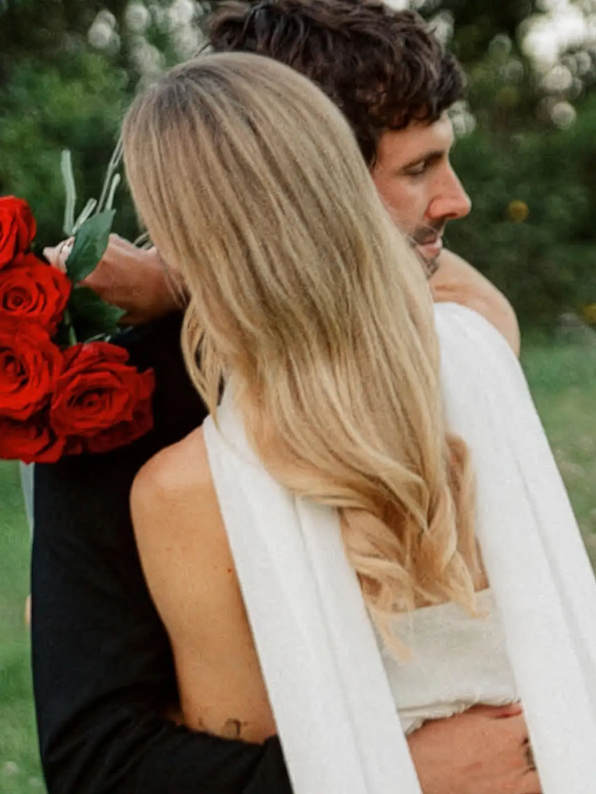 Bride's long, glossy blonde waves seen from behind as the groom holds red roses