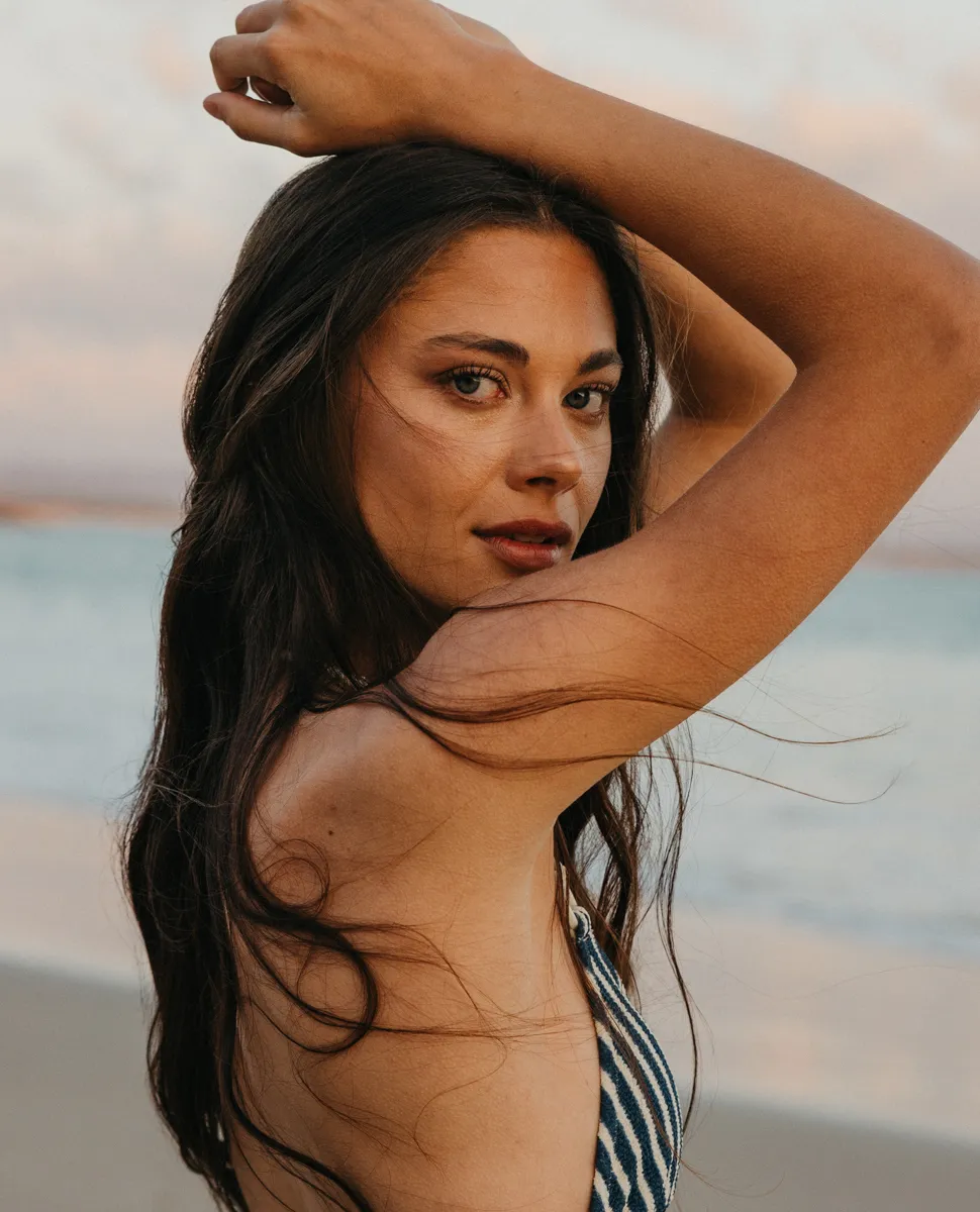 Woman with long dark hair and bronzed, natural makeup posing on the beach at sunset
